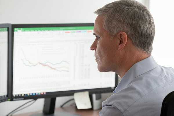 A man sits at a desk, looking at charts on his computer monitor.