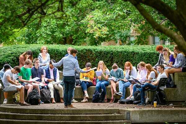 A class gathers outside, reading books. A professor stands before them.