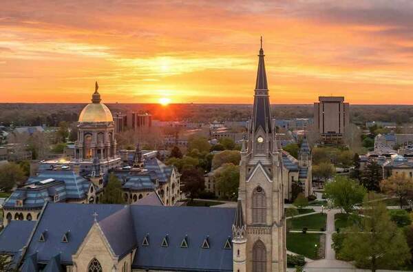 An East aerial view of Notre Dame Campus just as the sun is rising. The Basilica is rising at the front, with the main building and library in the background.
