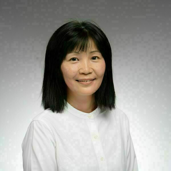An Asian woman with shoulder-length, straight dark hair and bangs smiles in front of a gray  gradient background. She is wearing a white top.