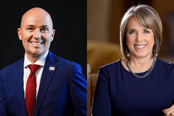 On the left is Spencer Cox, a smiling bald man in a navy blue suit, white shirt, and red tie. On the right is Michele Lujan Grisham, a smiling woman with short light brown hair wearing a navy blue top and a necklace.