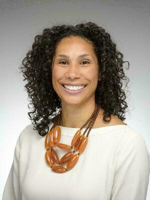 Anna Haskins, presenting as a woman with dark curly hair smiles, wearing a white top and a prominent amber-orange layered beaded necklace.