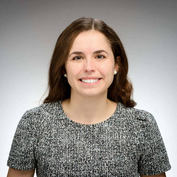 Jaclyn Biedronski is shown as a smiling young woman with long brown hair and pearl earrings. She wears a black, and white tweed top against a gray background.