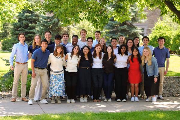 A group of approximately 20 smiling Notre Dame Poverty Research Fellow students, a diverse mix of men and women, standing together on a paved path outdoors. They wear various smart casual attire. Green trees and a brick building are visible in the sunny background.