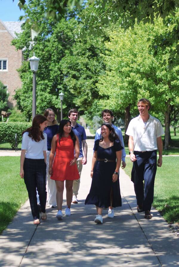 Seven smiling students walk on a sunny paved path on Notre Dame's campus. They wear diverse casual clothing as green trees and a stone campus building are visible behind them.