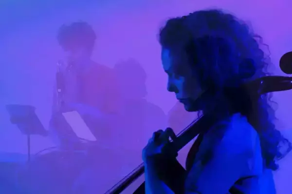 A person with dark, curly hair and a headset mic plays a cello, focused under vibrant blue and purple lighting. Another musician, indistinctly visible in the blurry background, plays a wind instrument.