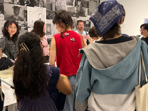 A group of students views an art exhibition. A guide explains works on a wall covered with black and white art. A student in glasses smiles; another wears a red top, and a person with a blue bandana wears a blue jacket.