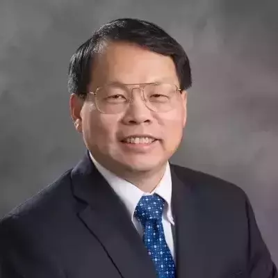 A smiling man with glasses in a dark suit, white shirt, and blue patterned tie against a grey background.