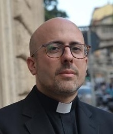 A bald man with a beard and glasses wears a black clerical shirt and white collar, looking directly forward.
