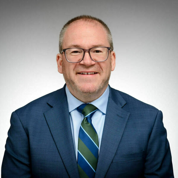 Smiling man wearing glasses, a blue suit, light blue shirt, and a striped blue and green tie.