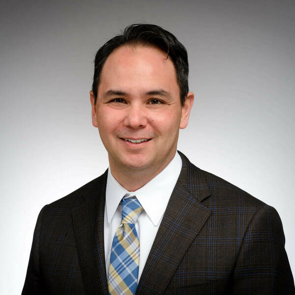 Man with dark hair, smiling in a brown suit jacket with blue plaid, white shirt, and blue and yellow plaid tie.