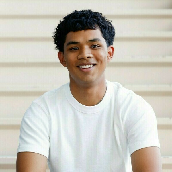Young man with dark, curly hair and a white t-shirt smiles brightly, against a light paneled background.