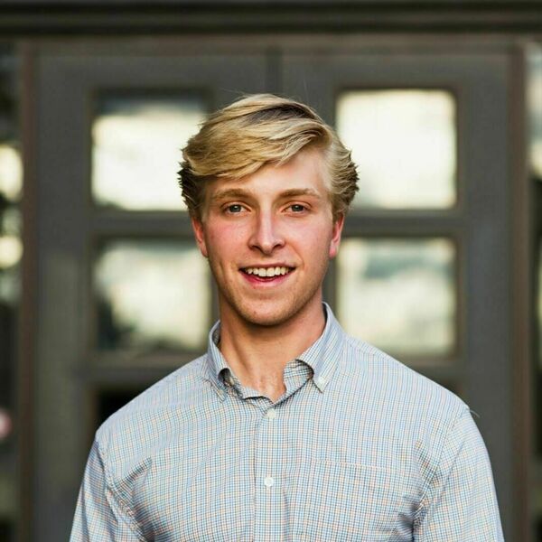 Young man with blond hair and a smile wears a blue and orange plaid shirt, standing before a blurry grey door.