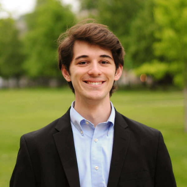 Smiling young man with brown hair in a light blue shirt and dark blazer, standing outdoors.