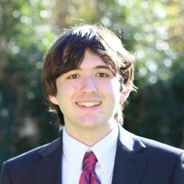 Smiling young man with dark hair wearing a suit, striped shirt, and burgundy tie, standing outdoors.