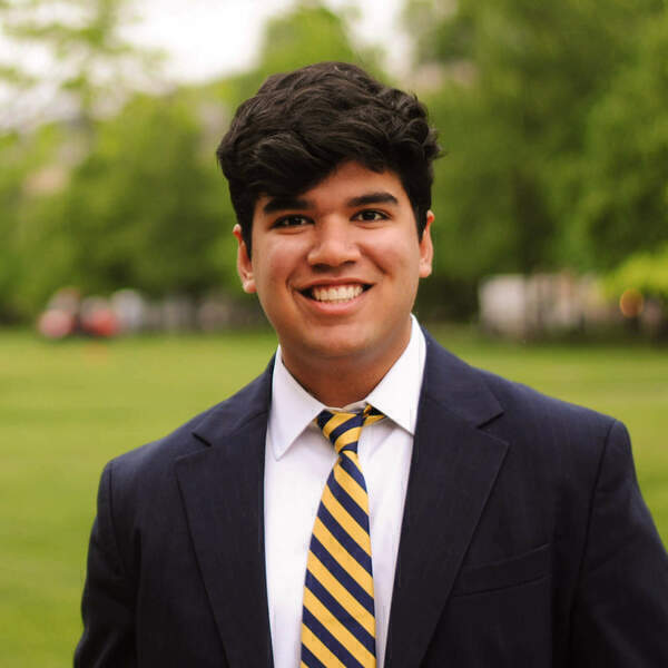 A smiling young man in a dark blue blazer, white shirt, and yellow and blue striped tie stands outdoors.