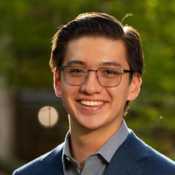 A young man with dark hair and glasses smiles broadly. He wears a blue blazer and light blue shirt.