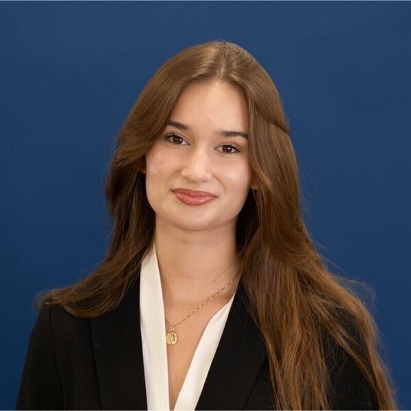 Young woman with long brown hair, wearing a black blazer and white blouse, smiles faintly against a blue background.
