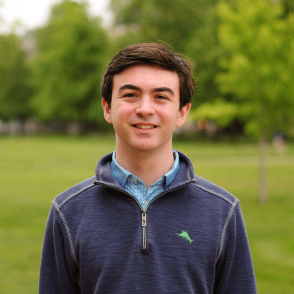 Young man with dark hair wears a blue quarter-zip over plaid shirt, smiling outdoors with green trees and grass.