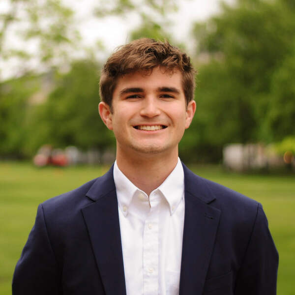 Young man with brown hair and a broad smile, wearing a navy blue blazer and white shirt, standing outside.