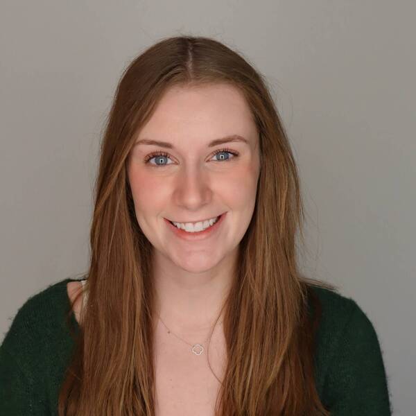 A smiling young woman with long brown hair and blue eyes wears a green V-neck sweater over a white lace top.