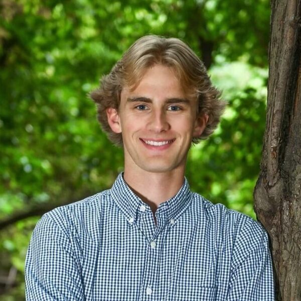 Young man with blond hair, blue eyes, and a blue gingham shirt smiles next to a tree with green foliage.