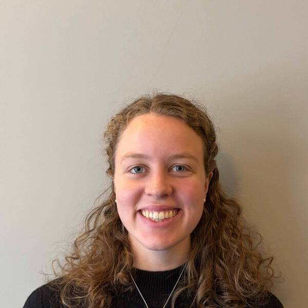 A young woman with curly brown hair and blue eyes smiles, wearing a black ribbed sweater and a silver necklace.