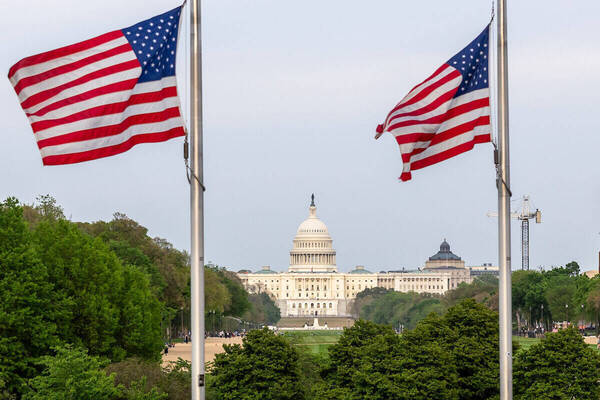 Two US flags wave on poles with the white-domed US Capitol Building centered in the background surrounded by green trees.