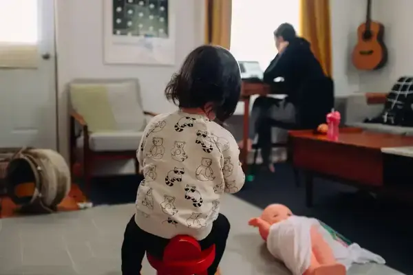 Toddler in a white bear-and-raccoon shirt on a red stool faces an adult working on a laptop.