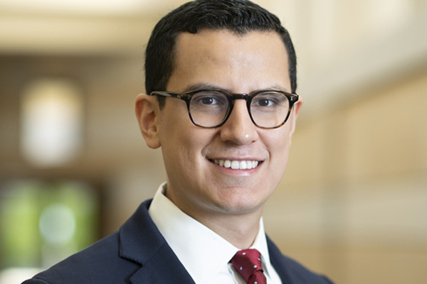 A smiling man with dark hair, tortoise-shell glasses, a navy suit jacket, white shirt, and a red polka-dot tie.