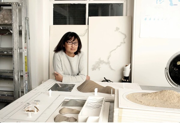Maya Lin photo. Woman in glasses and a grey sweater sits with arms crossed behind detailed architectural landscape models, looking at viewer.