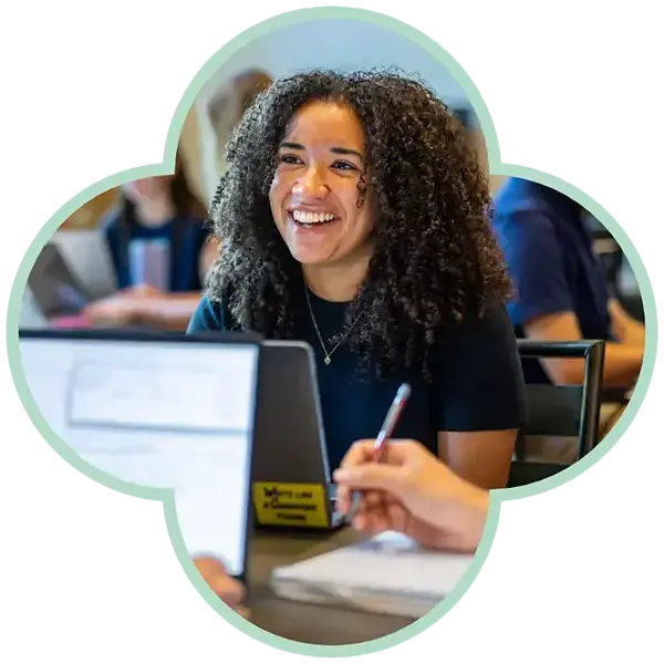 A smiling woman with curly dark hair wears a black shirt and looks to the side in a classroom setting.